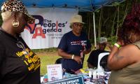 A woman stands behind a table with voting rights signage and speaks to two other women standing in front of her.