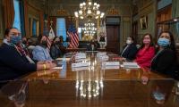 Women wearing Covid masks sitting around a large wooden table with American flags in the background.