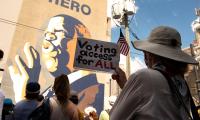 A woman with her back to the camera holds a sign which says "Voting access for ALL", while standing with others in front of a large mural of John Lewis.