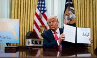 Donald Trump sitting at a desk in the Oval Office holding up an executive order with his signature on it.