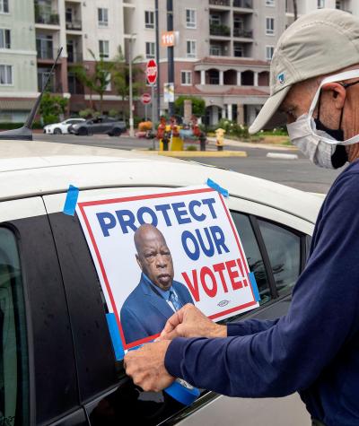 A man wearing a mask tapes a sign to the window of a car. The sign reads "Protect our vote!" and has a picture of John Lewis.