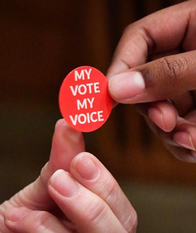 Close up of a person handing a red sticker which says "My Vote, My Voice" to another person
